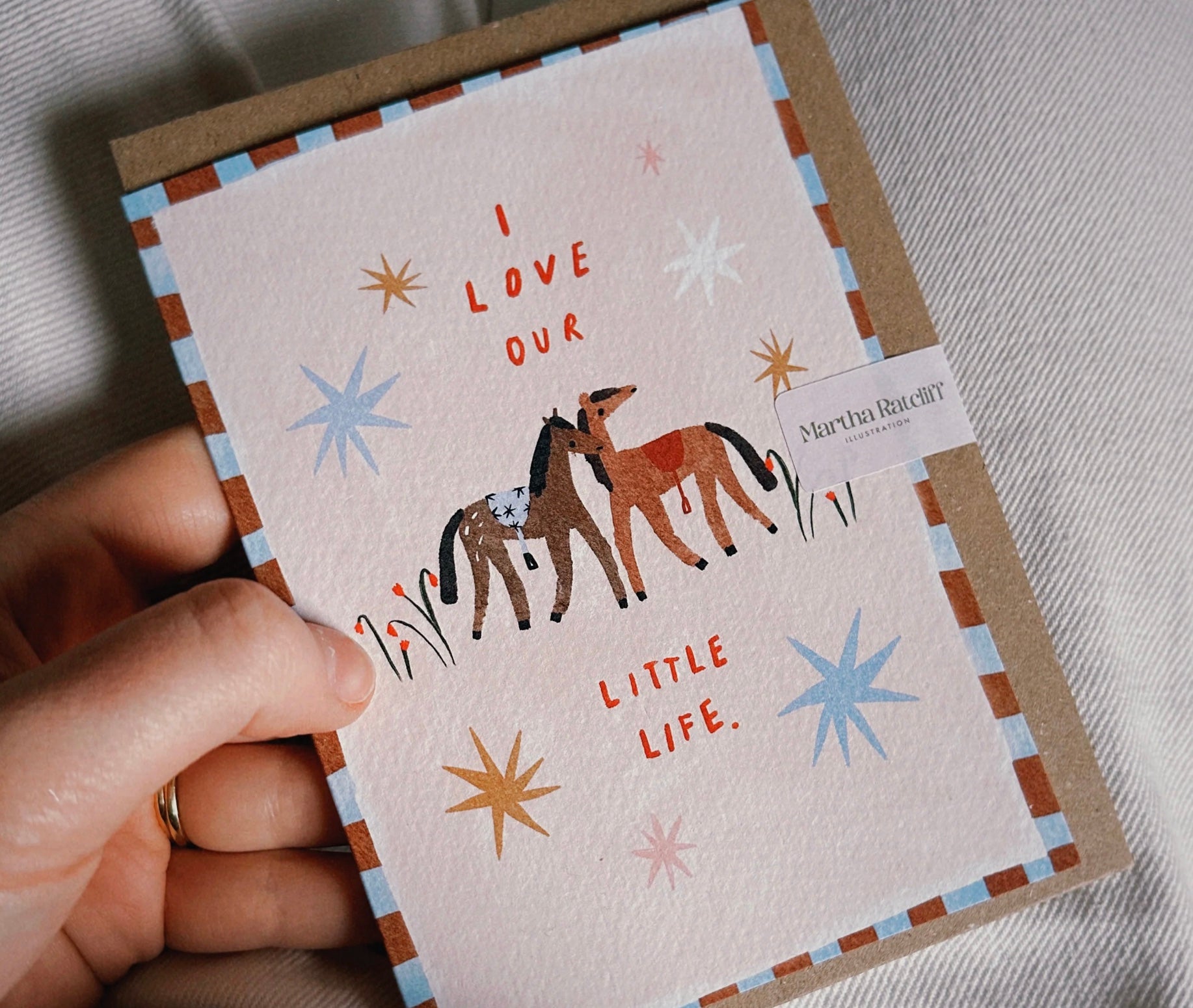 A hand holds a square greeting card with a kraft envelope behind it. The card has a cream background with a brown and blue striped border, an illustration of two horses, and the text 'I LOVE OUR LITTLE LIFE.