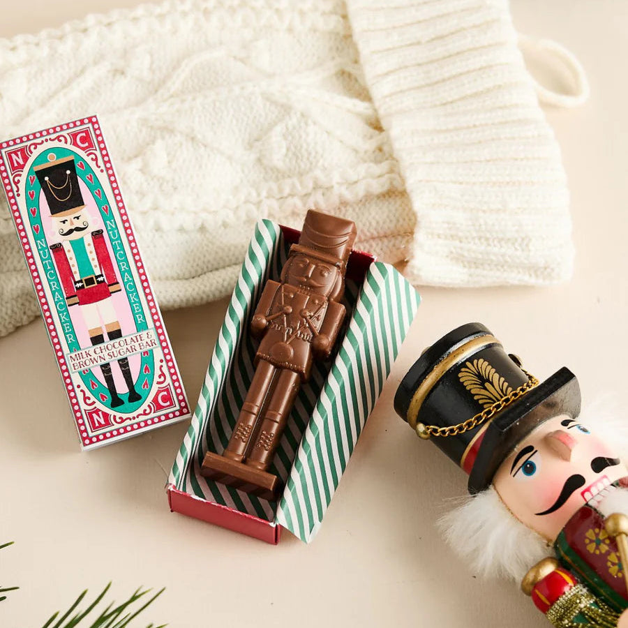 Chocolate nutcracker in a striped box with a decorative card, surrounded by Christmas decorations on a light background.