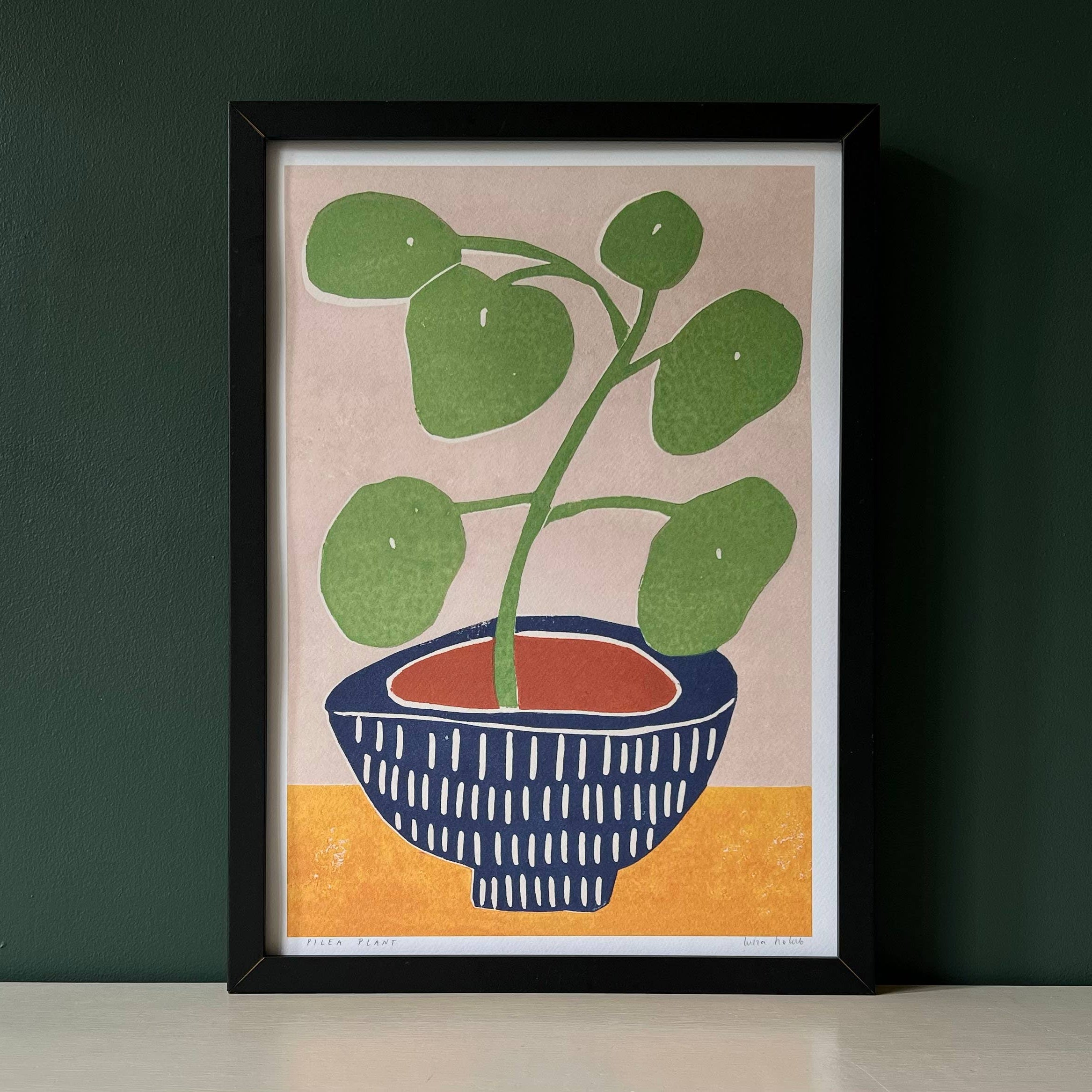 Framed artwork of a potted plant on a beige background with a dark green wall.